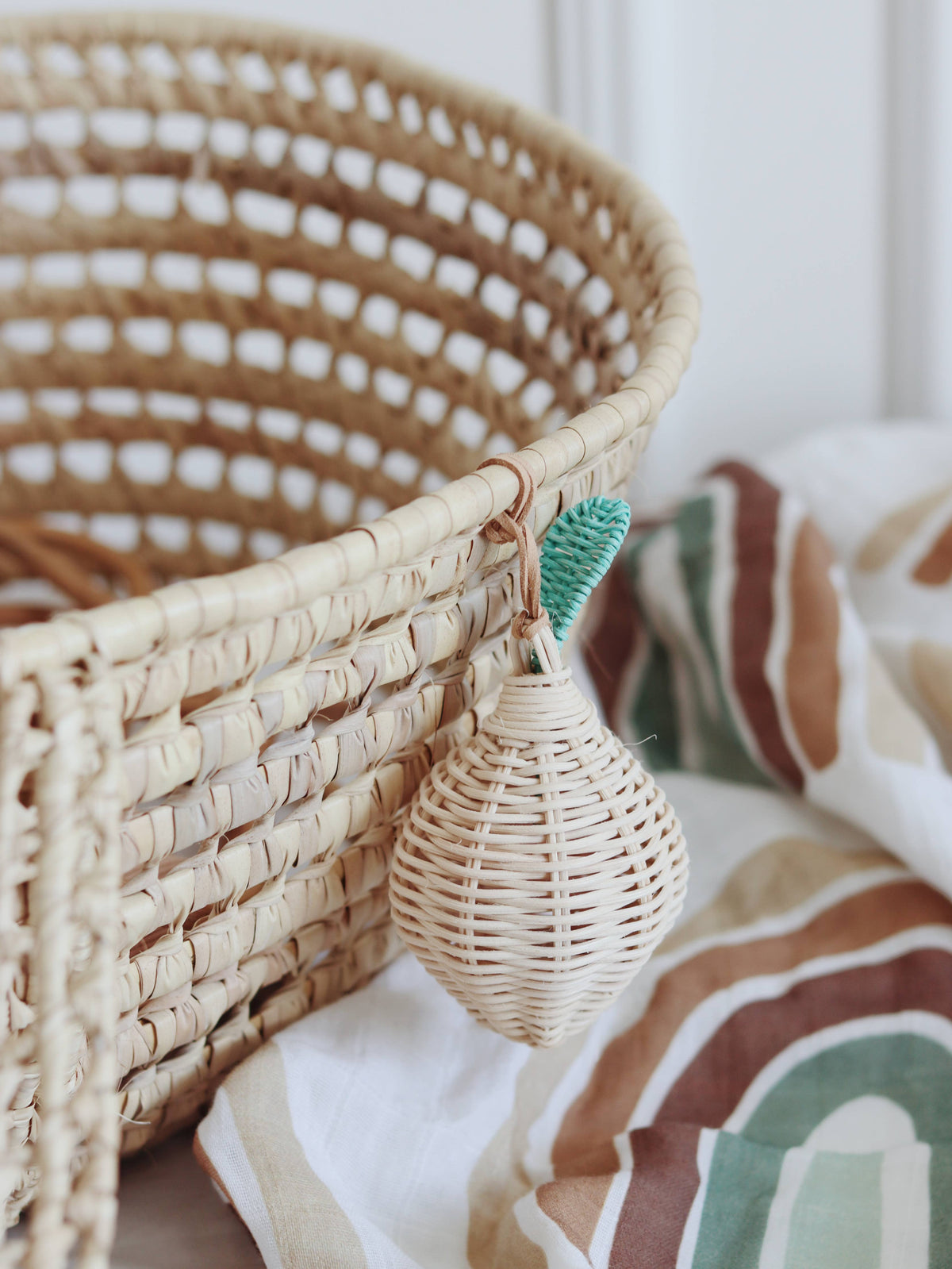 Handmade woven rattan lemon-shaped rattle with a green leaf detail and tan leather string, hanging from the edge of a natural rattan basket.
