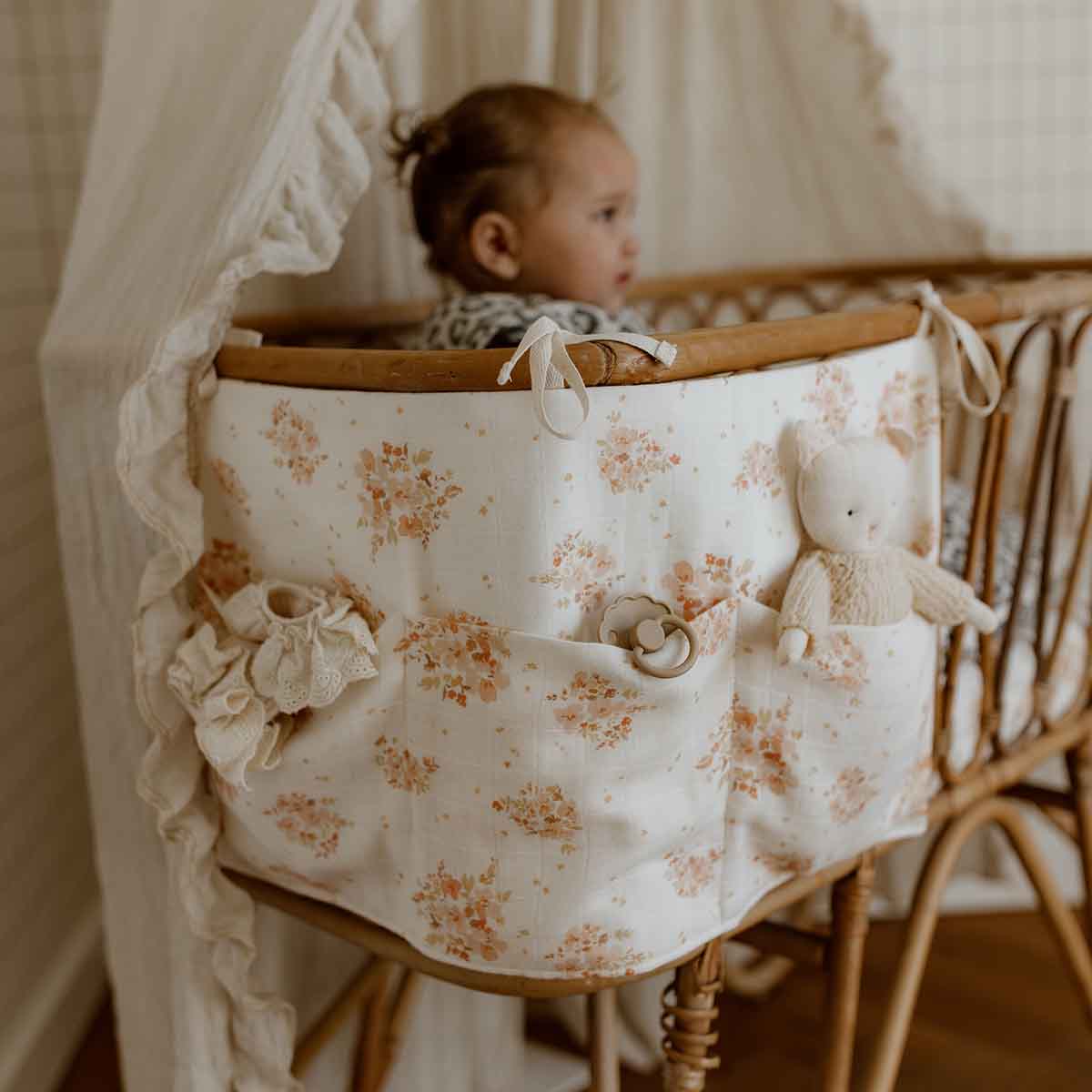 Mathilde Bouquet crib organiser in cream cotton with a delicate peach floral print, attached to a wooden baby crib with fabric ties and front pockets.