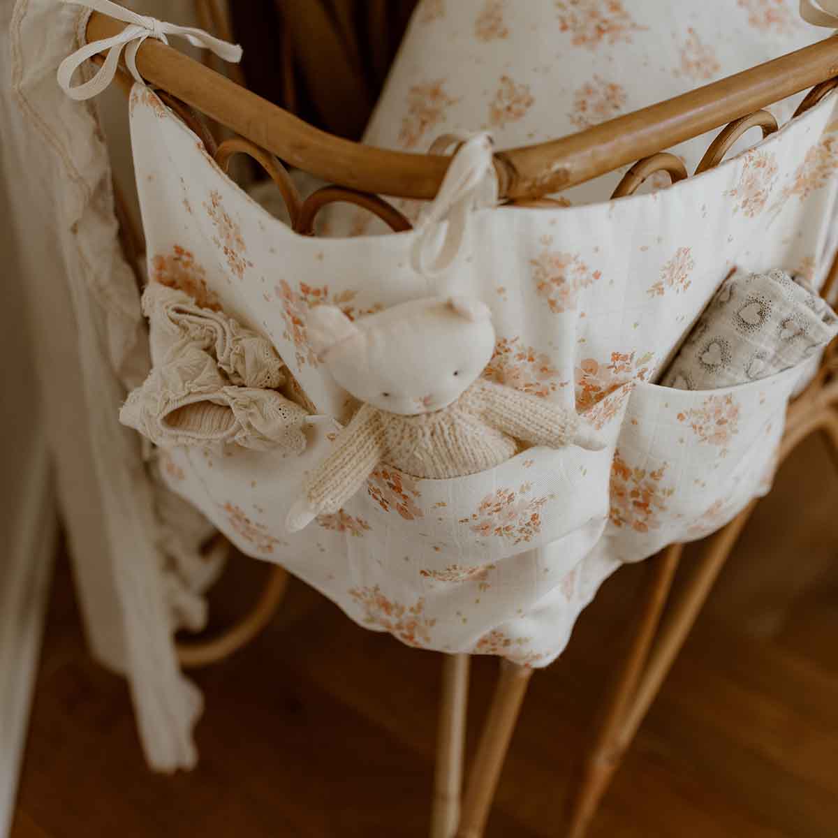 Mathilde Bouquet crib organiser in cream cotton with a peach floral print and front pockets, attached to a wooden crib and styled with a knitted bunny and baby blanket.