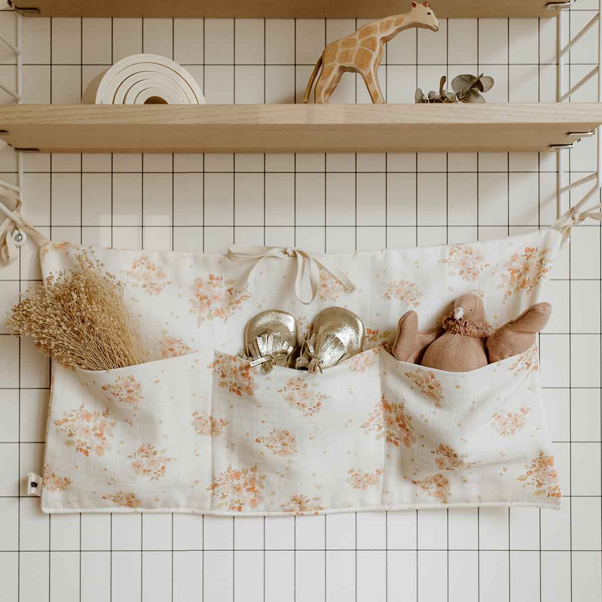 Mathilde Bouquet organiser in cream with a peach floral print and multiple storage pockets, hung on a wire grid wall and holding baby accessories, a dried flower arrangement and a soft toy.