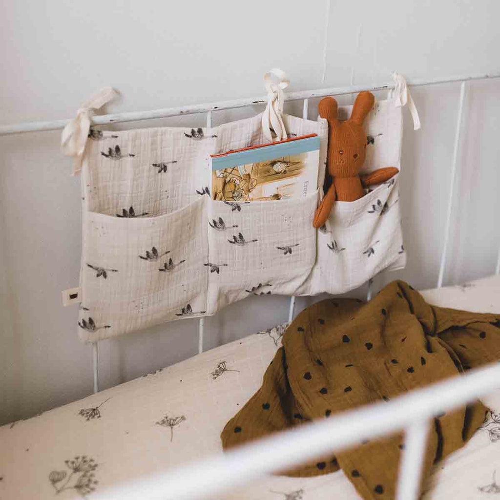 Mathilde Goose Natural cot storage organiser hanging from a white cot rail, with a book and brown rabbit soft toy tucked into its pockets above mustard-brown patterned bedding.