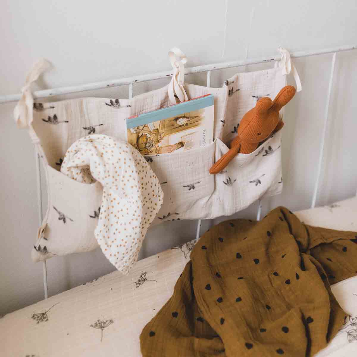 Natural cotton cot storage organiser with a grey goose print, attached to a cot rail and holding a muslin cloth, a children's book and a brown rabbit soft toy.