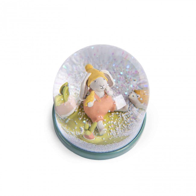 Moulin Roty Three Little Rabbits snow globe with a rabbit and friends in a garden scene surrounded by falling snowflakes on a white background