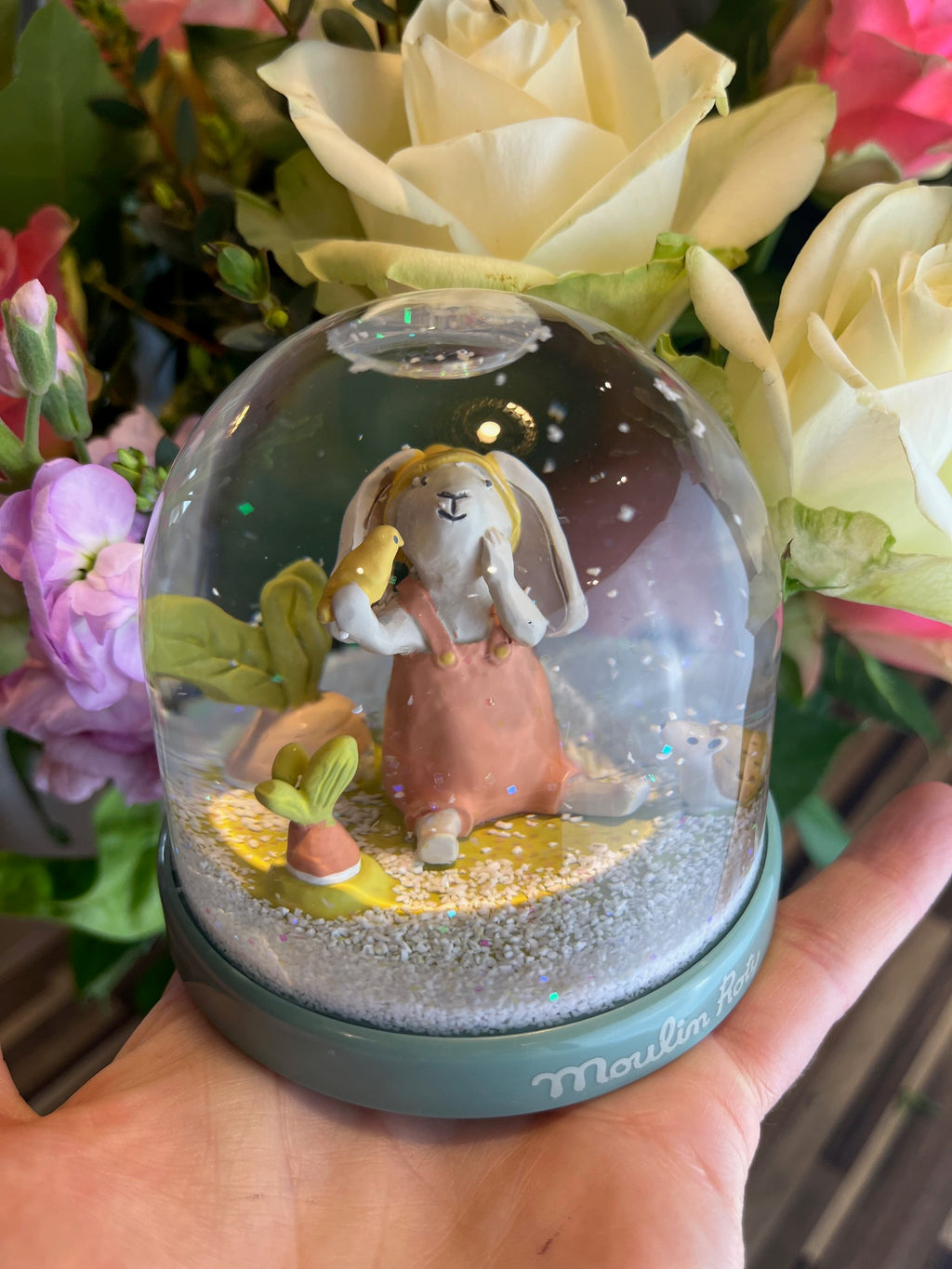 Moulin Roty Three Little Rabbits snow globe featuring a rabbit in a garden scene with flowers and falling snowflakes