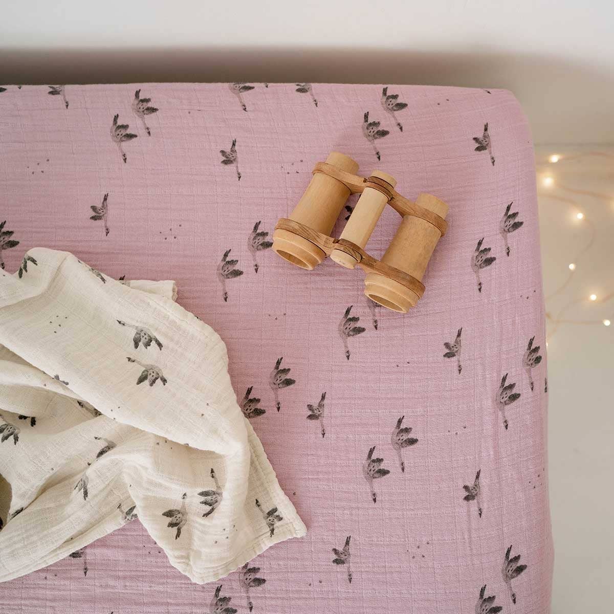 Lilac fitted cot sheet with a delicate grey goose print displayed on a mattress alongside a matching cream muslin cloth and wooden toy binoculars.