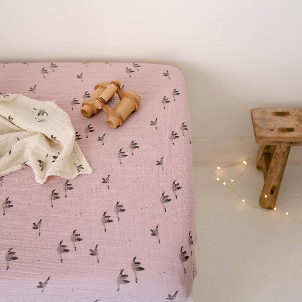 Lilac goose-print fitted cot sheet styled on a mattress with a matching muslin cloth and wooden toy binoculars in a softly lit nursery.