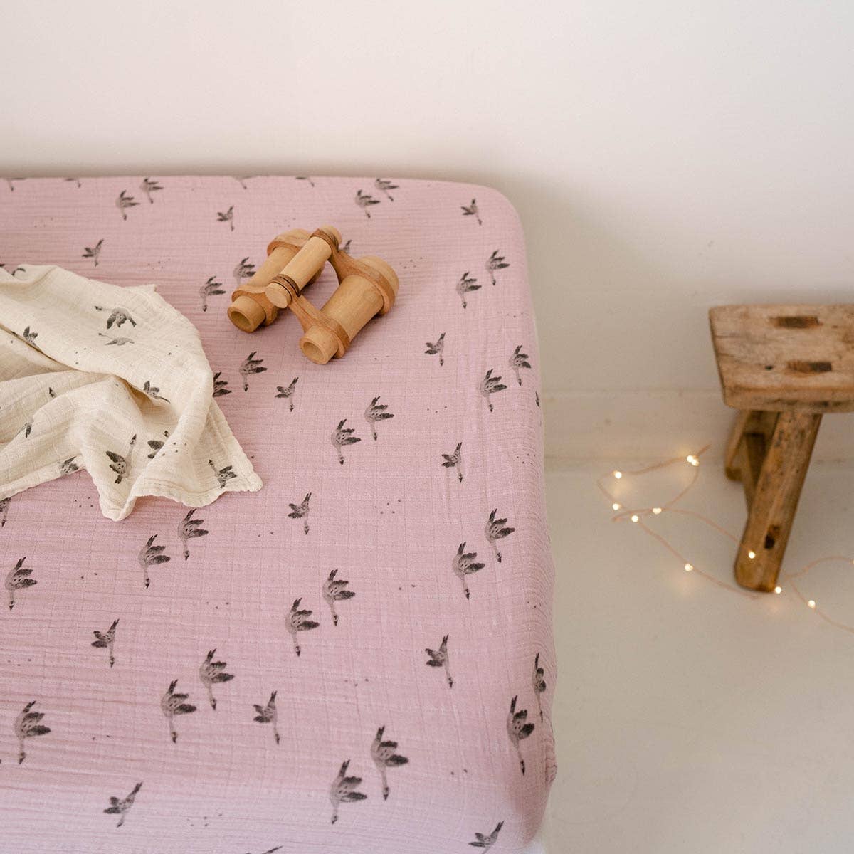 Lilac goose-print fitted cot sheet styled on a mattress with a matching muslin cloth and wooden toy binoculars in a softly lit nursery.