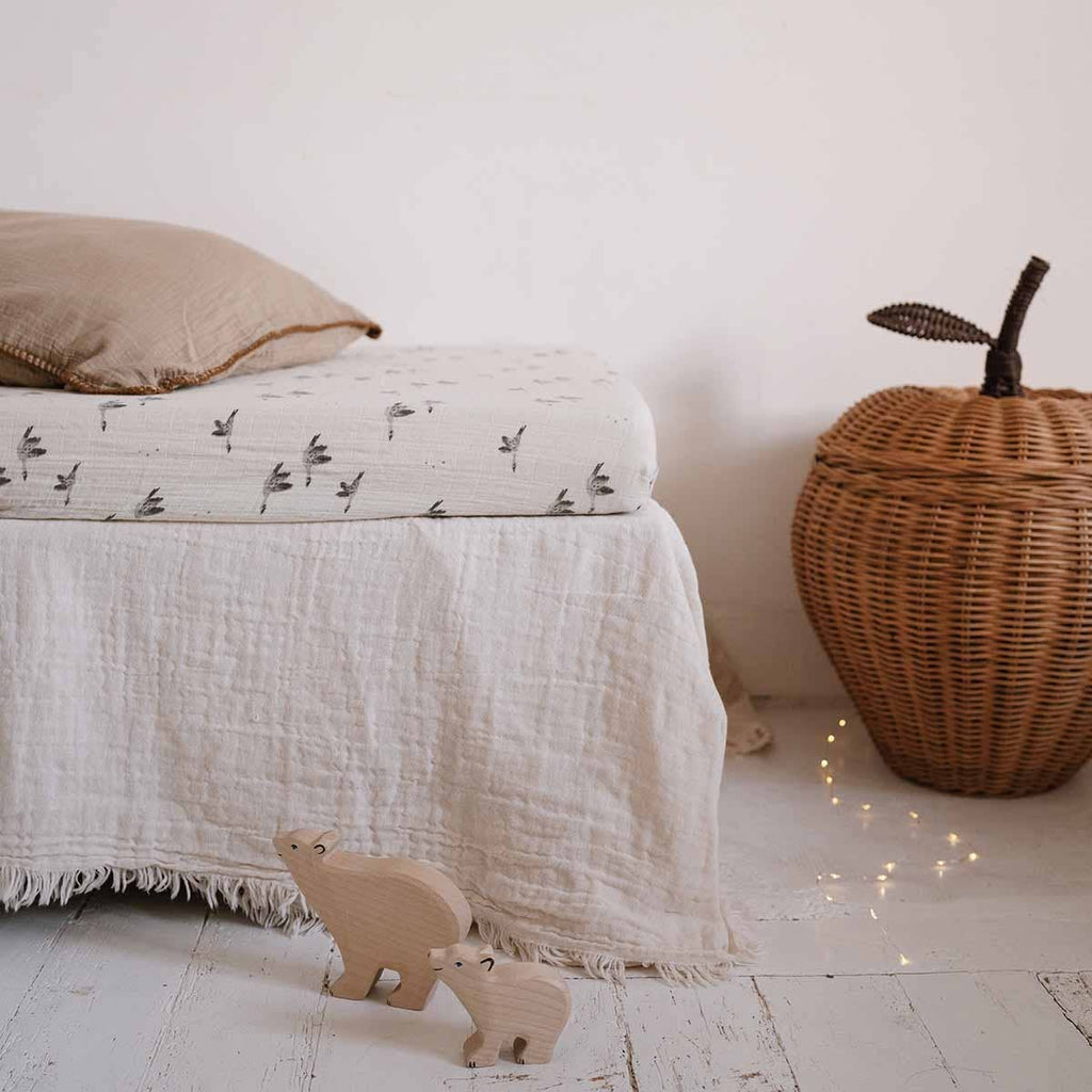Olivia Goose Natural fitted cot sheet with a delicate grey goose print, styled on a cot with neutral bedding, a woven apple basket and wooden animal toys.