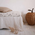 Olivia Goose Natural fitted cot sheet with a delicate grey goose print, styled on a cot with neutral bedding, a woven apple basket and wooden animal toys.
