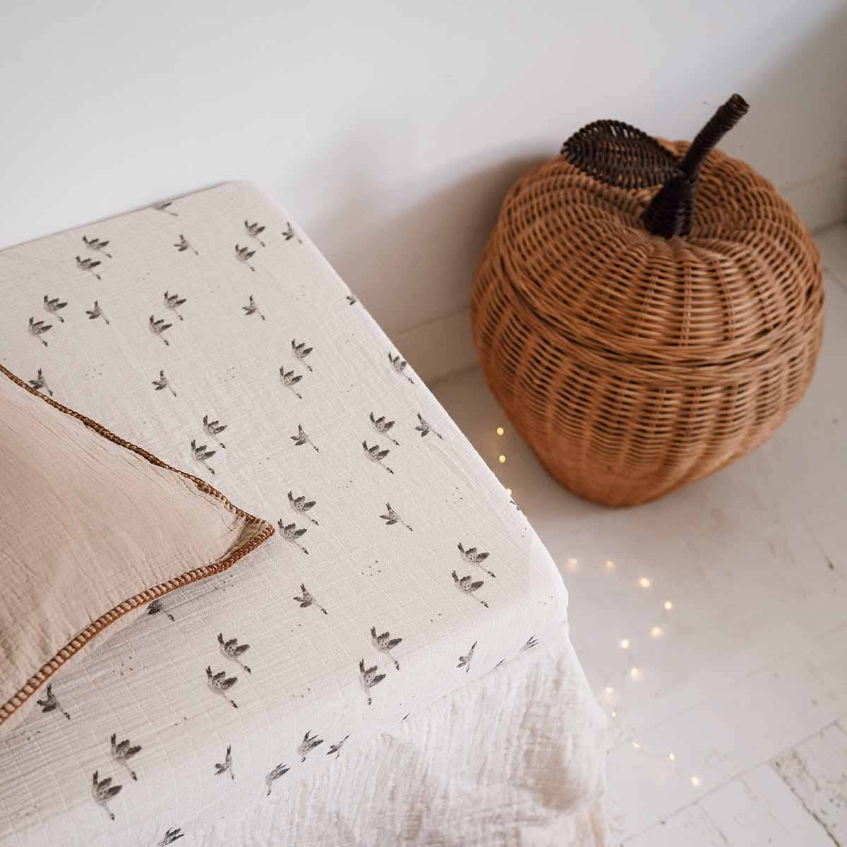 Olivia Goose Natural fitted cot sheet featuring a delicate grey goose print, styled with a beige cushion and a woven apple-shaped basket.