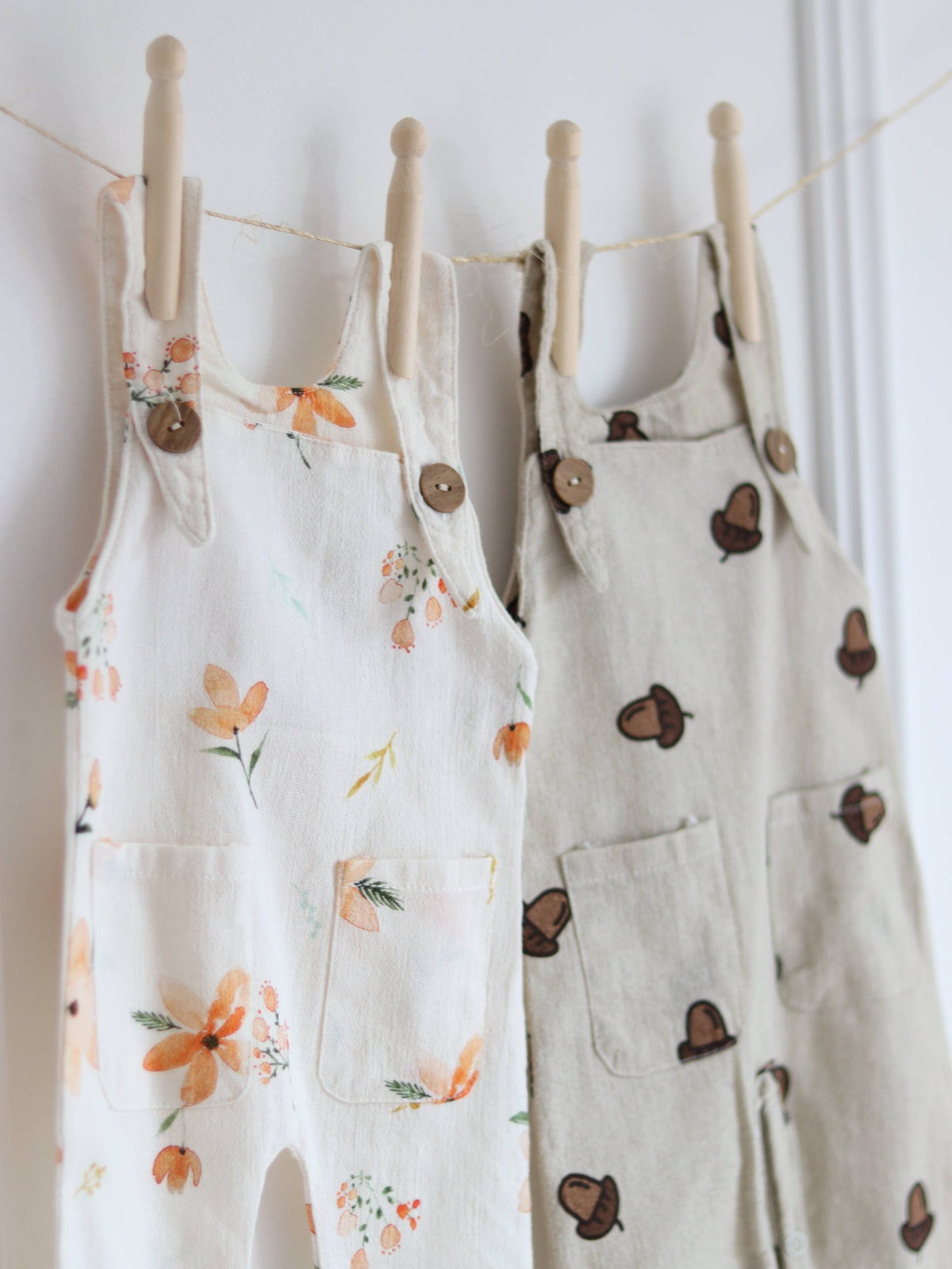 Two baby dungarees hanging side by side, featuring a cream floral print in peach and orange and a beige acorn print, both with wooden-look shoulder buttons and front pockets.