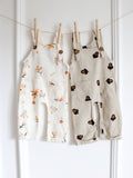 Two baby dungarees hanging side by side, one in cream with a delicate floral print and the other in beige with a brown acorn print, both featuring shoulder straps with wooden-look buttons.