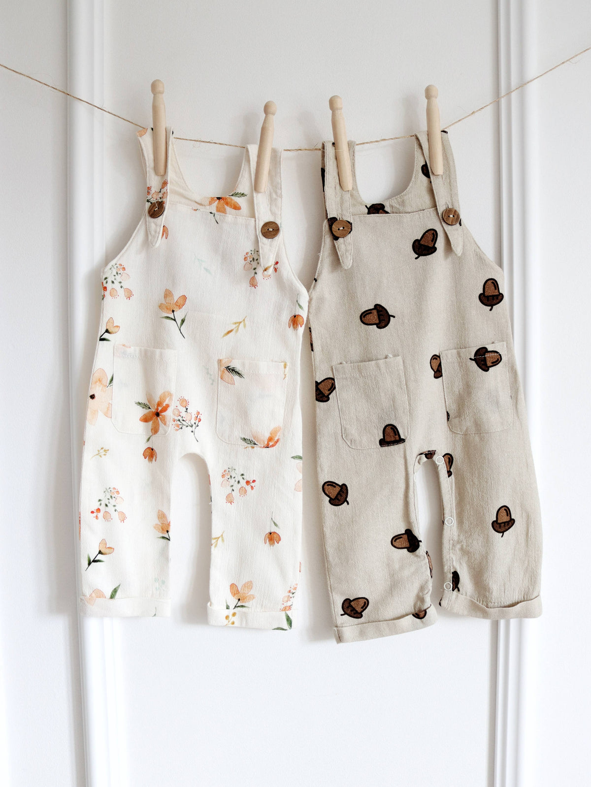 Two baby dungarees hanging side by side, one in cream with a delicate floral print and the other in beige with a brown acorn print, both featuring shoulder straps with wooden-look buttons.