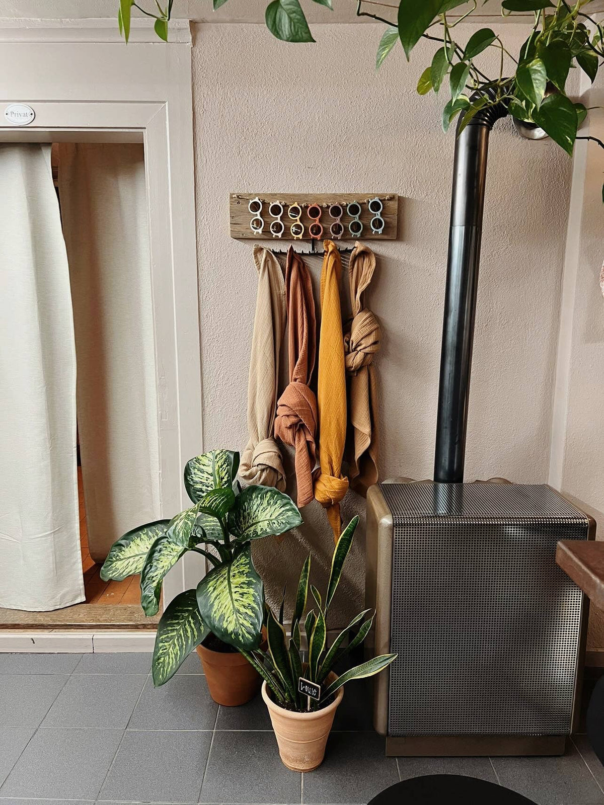 Neutral-coloured muslin blankets in cinnamon, beige, mustard and camel hanging on a wooden wall rack beside potted plants and a wood-burning stove.