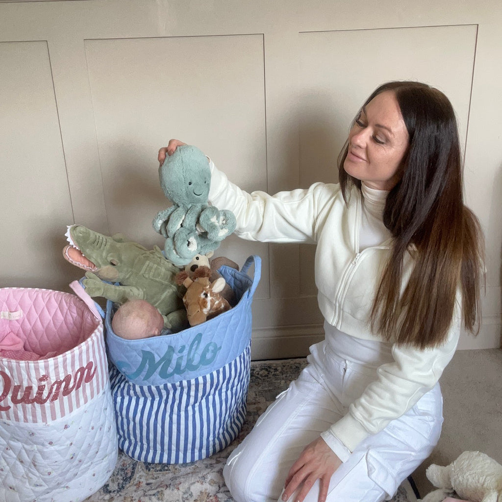Two quilted toy storage bags in pink and blue stripe designs filled with soft toys, with a woman organising a blue octopus toy in the blue bag.