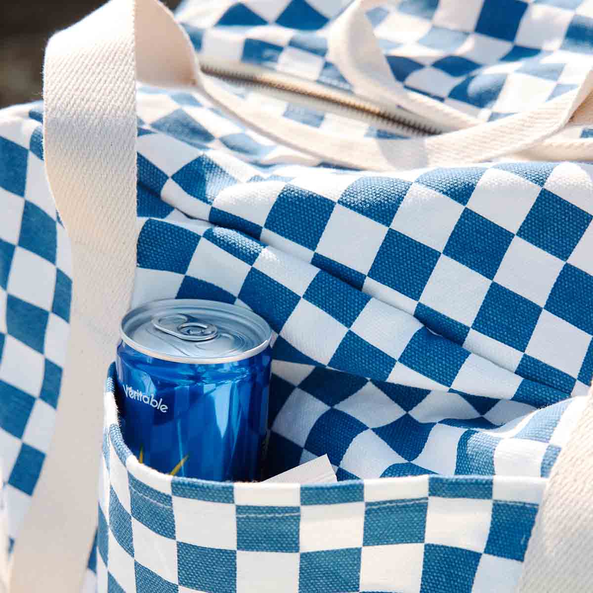 Close-up of the Raphael Checkerboard Blue bag's exterior pocket holding a blue drink can, showing the cotton canvas checkerboard pattern and cream h