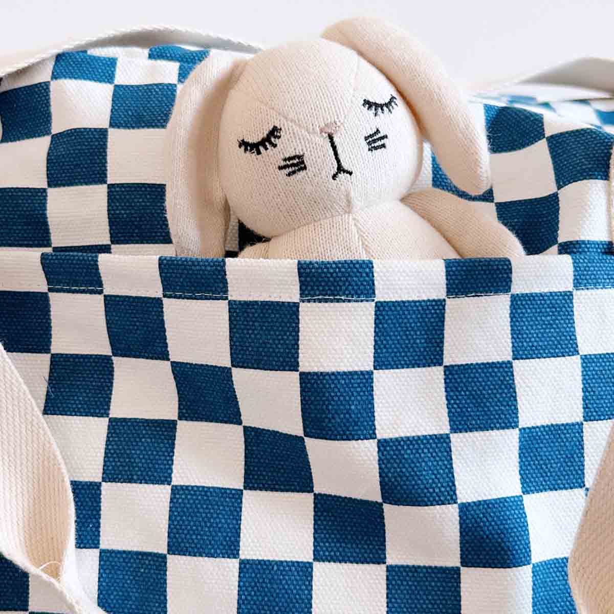 Close-up of a cream knitted rabbit soft toy peeking out of the front pocket of the navy blue and ecru checkerboard Raphael nappy bag.