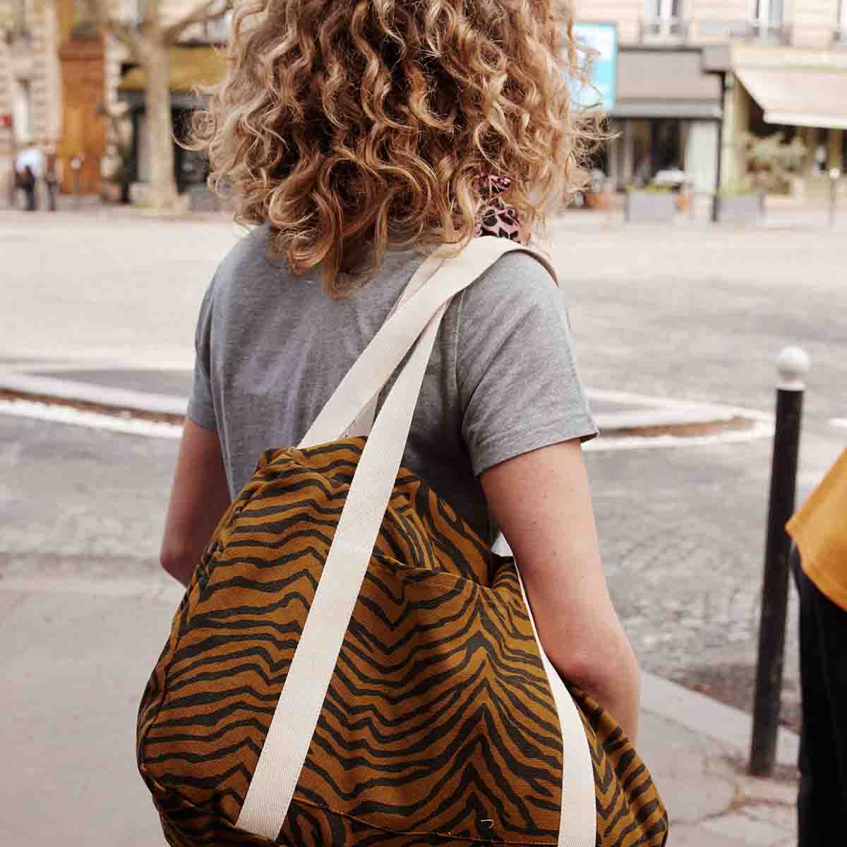 Rear view of a woman carrying the Tapenade Zebra Raphael weekend bag, showing the brown and black striped pattern and long cream shoulder straps.