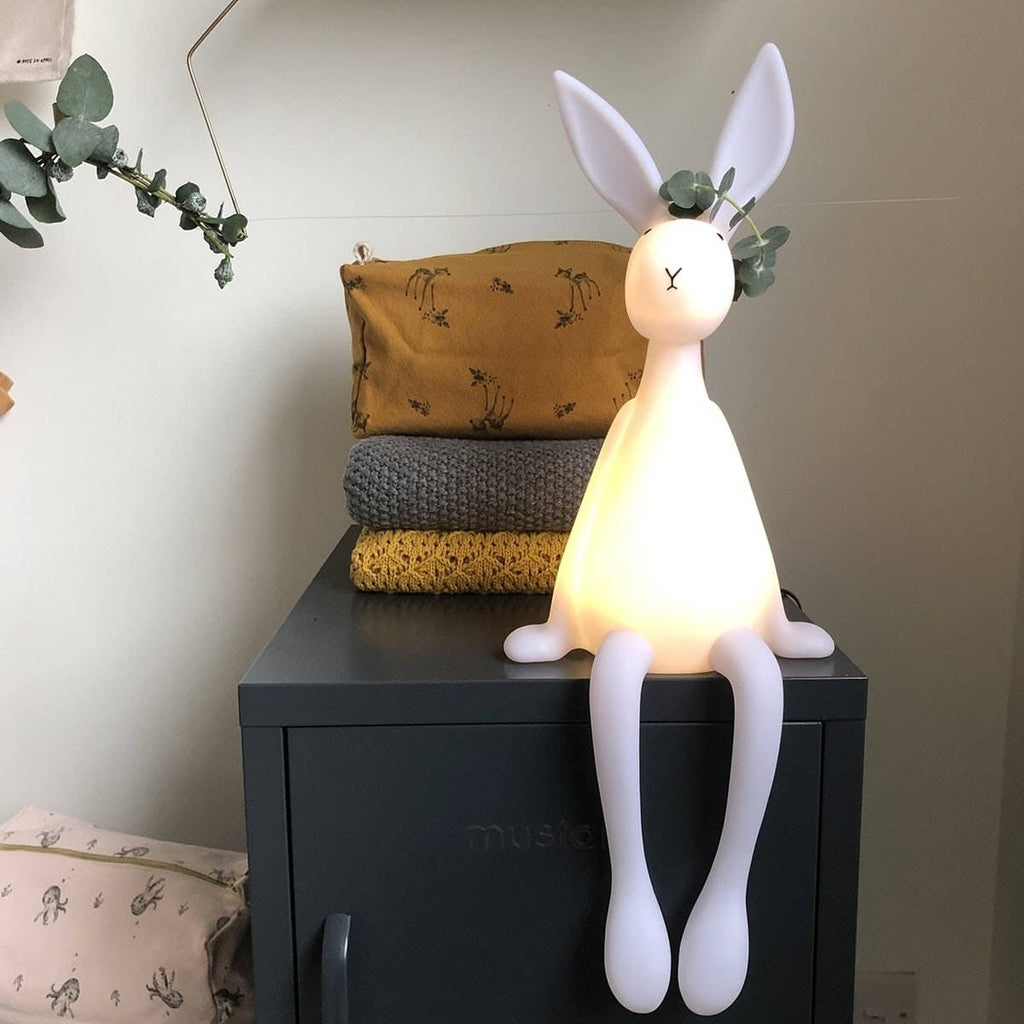 Joseph Bunny white LED night light with long ears and dangling legs, glowing warmly while seated on a dark bedside cabinet in a neutral nursery.
