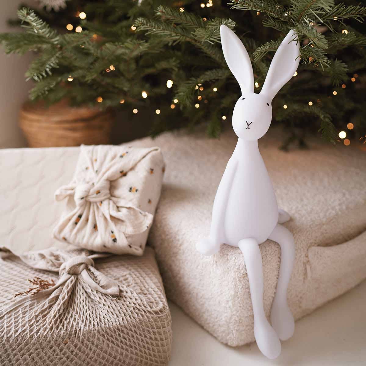 White Joseph Bunny night light styled beside wrapped gifts and cream knitted blankets under a Christmas tree with warm fairy lights.