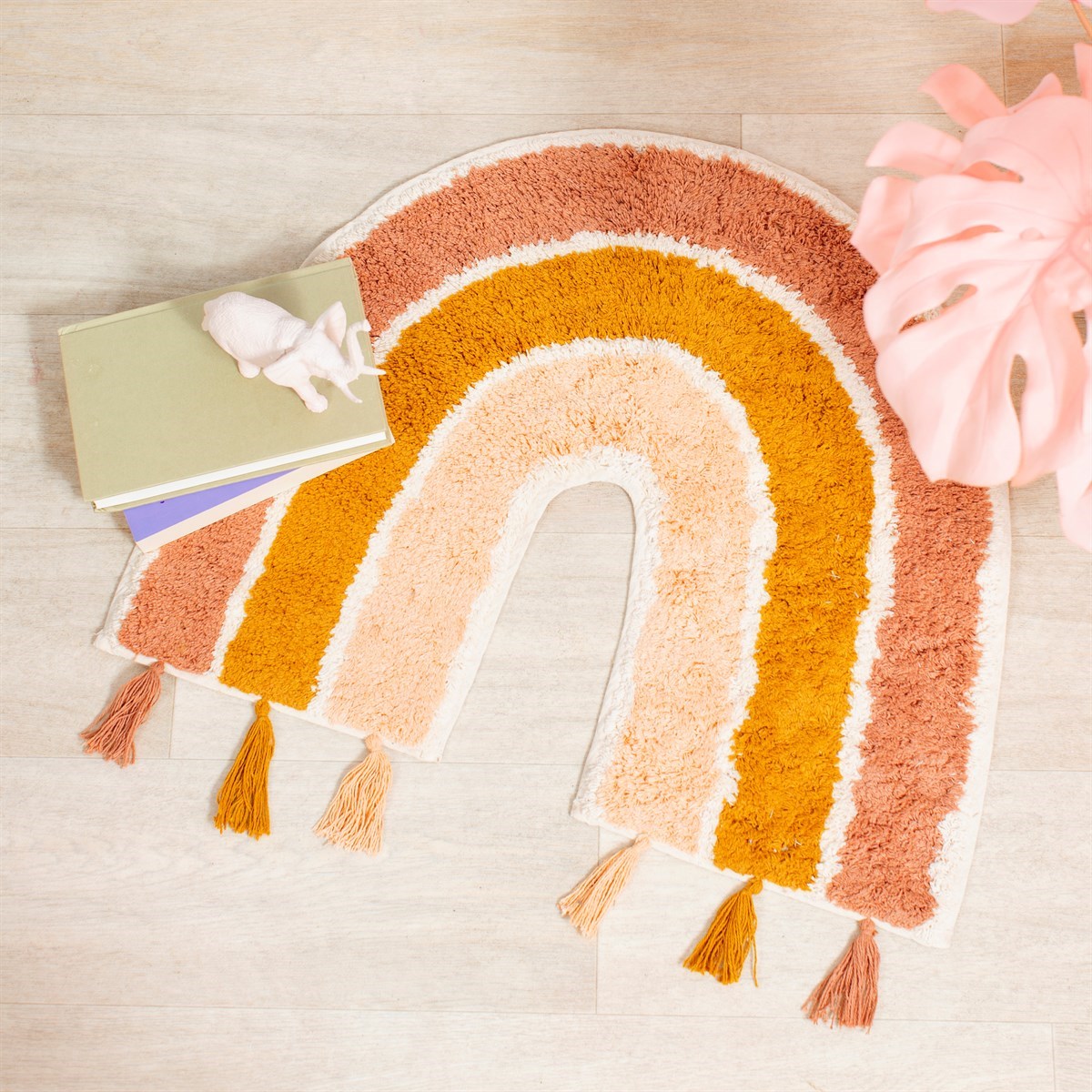 Earth Rainbow rug by Sass & Belle in terracotta pink, mustard yellow and cream, styled on a light wood floor with books, a small toy and pink decorative foliage.