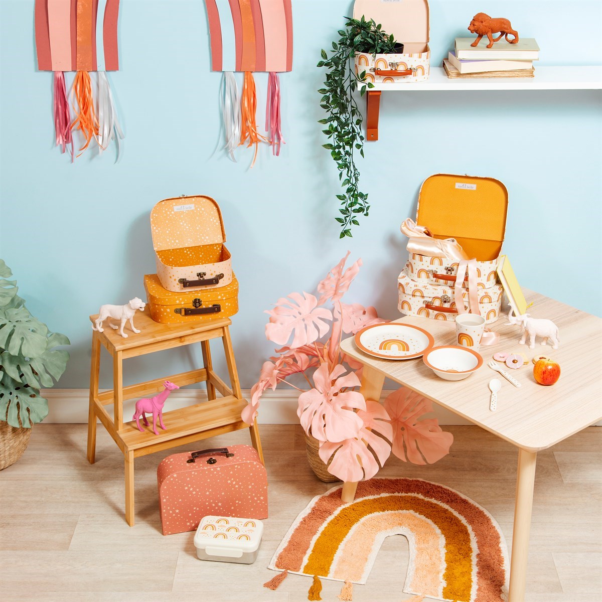 Sass & Belle Earth Rainbow rug in terracotta pink, mustard yellow and cream, styled in a colourful children’s playroom with a wooden table, toy suitcases, animal figures and rainbow accessories.
