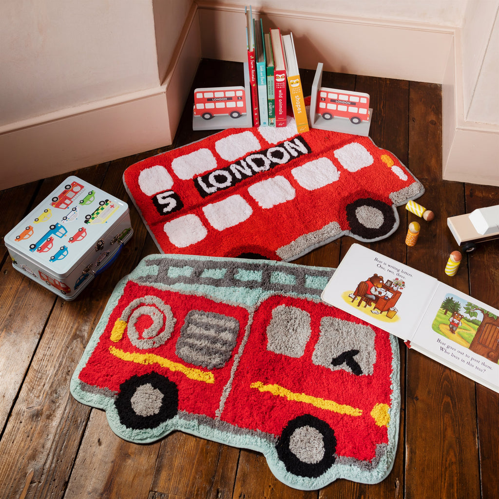 Red fire engine and London bus rugs displayed on a dark wooden floor with transport-themed books, toy vehicles and a children's suitcase.
