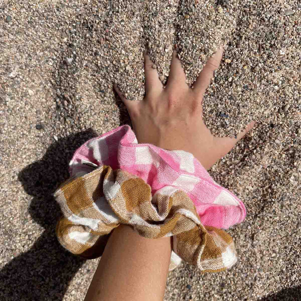 Caramel and cream gingham scrunchie worn on a wrist alongside a pink and cream checked scrunchie, photographed outdoors on a sandy surface.