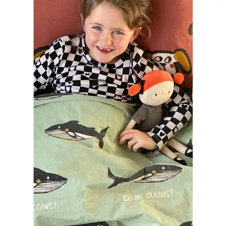 Child sitting in bed under sage green whale-print bedding, holding a soft toy and showing the whale illustrations and Clean Oceans message.