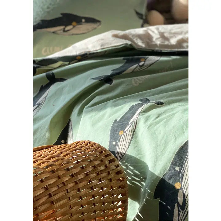 Close-up of sage green bedding featuring repeating dark whale illustrations with small yellow details, styled beside a woven rattan basket.