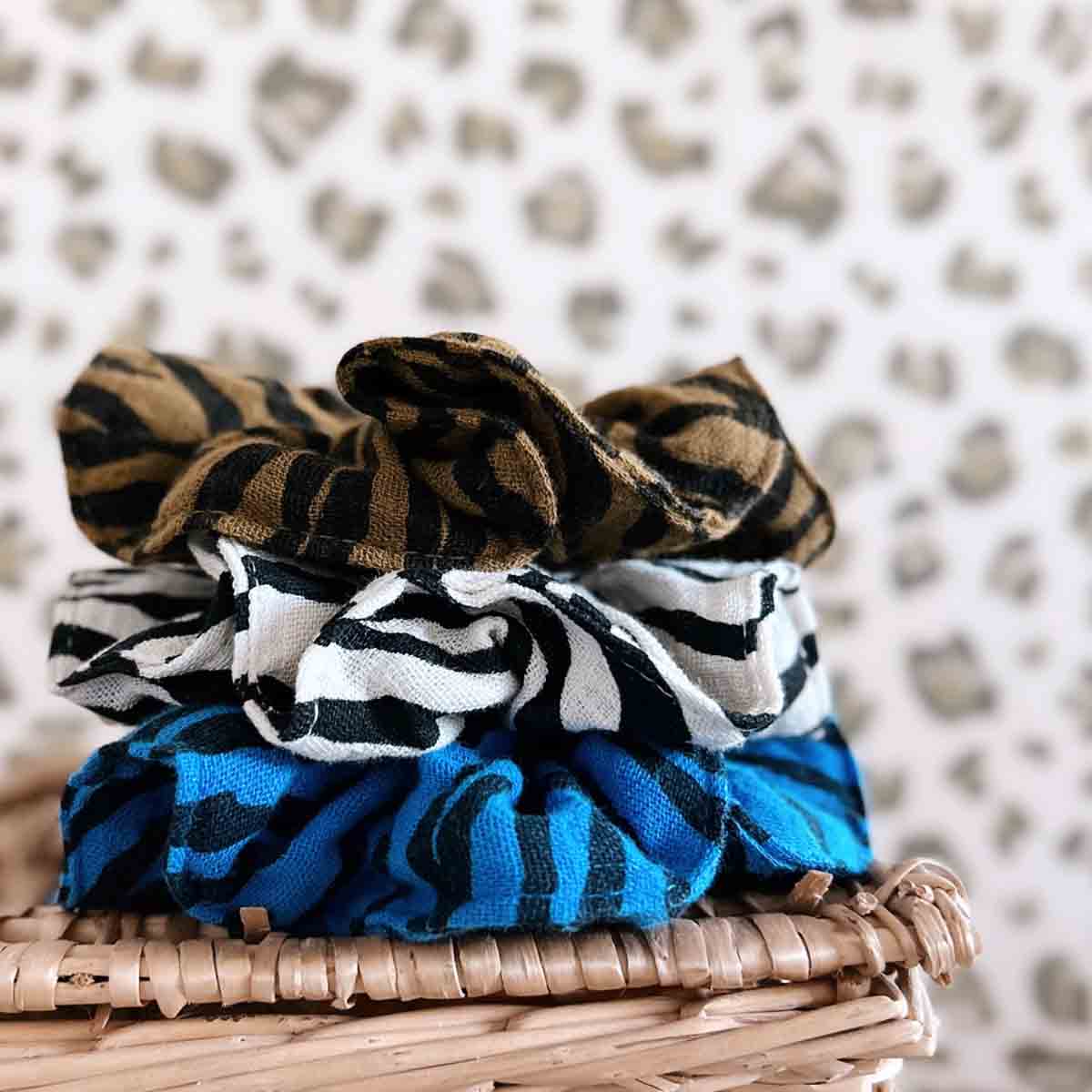 Three cotton muslin scrunchies in blue zebra, black and white zebra, and brown animal prints stacked on a woven basket.